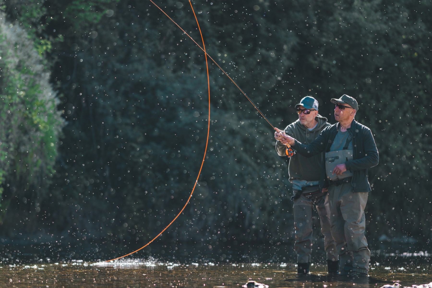 Zwei Fliegenfischer im Fluss üben gemeinsam den Wurf – orange Schnur tanzt über dem Wasser im Schweizer Auengewässer mit Flyfishing Guide Service. Flyfishing Guide Service, Flyfish Bünti, Fliegenfischen mit Rolf Bünter, Fliegenfischen Schweiz, Wurfübung Fluss, orange Flugschnur, Tandem-Wurf, Schweizer Auengewässer, Wassertropfen Wurf, Wurftechnik Coaching, Wathose, Brustpack, Fliessgewässer-Guiding, Stammrevier Töss, Hand-an-Hand-Wurf, Fliegenfischer-Coach, Wurfschule
