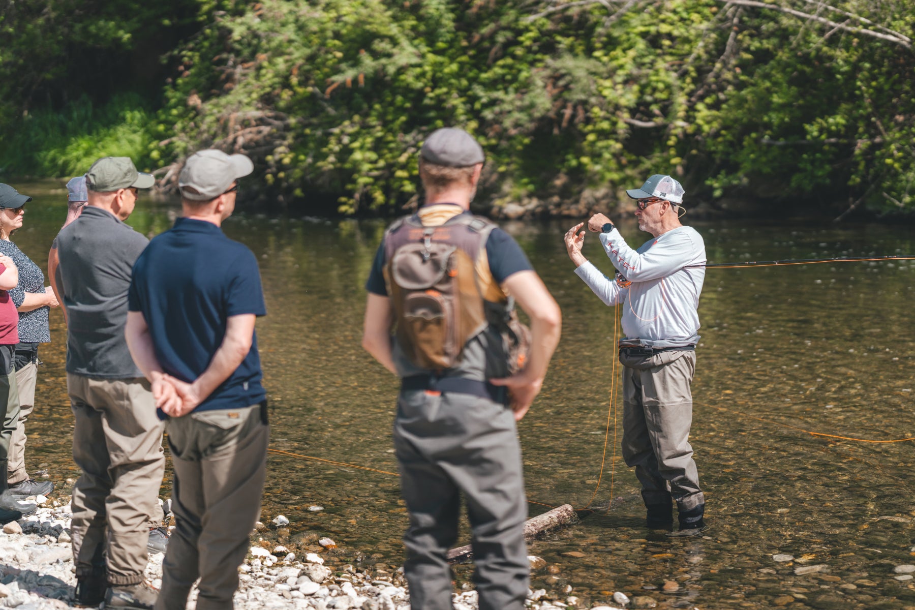 Rolf Bünter erklärt einer Anfängergruppe im Fluss den Aufbau der Fliegenmontage – Theorie am Wasser im Anfängerkurs von Flyfishing Guide Service. Flyfishing Guide Service, Flyfish Bünti, Fliegenfischen mit Rolf Bünter, Fliegenfischen Schweiz, Theorie am Wasser, Fliegenmontage erklären, Anfängergruppe, Wurfweste, Brustpack, Fliegenfischer-Theorie, Kursgruppe Fluss, Schweizer Auengewässer, Stammrevier Töss, Lehrmoment, Vorfach erklären, Knotenkunde, Coaching Praxis, Lernen am Wasser
