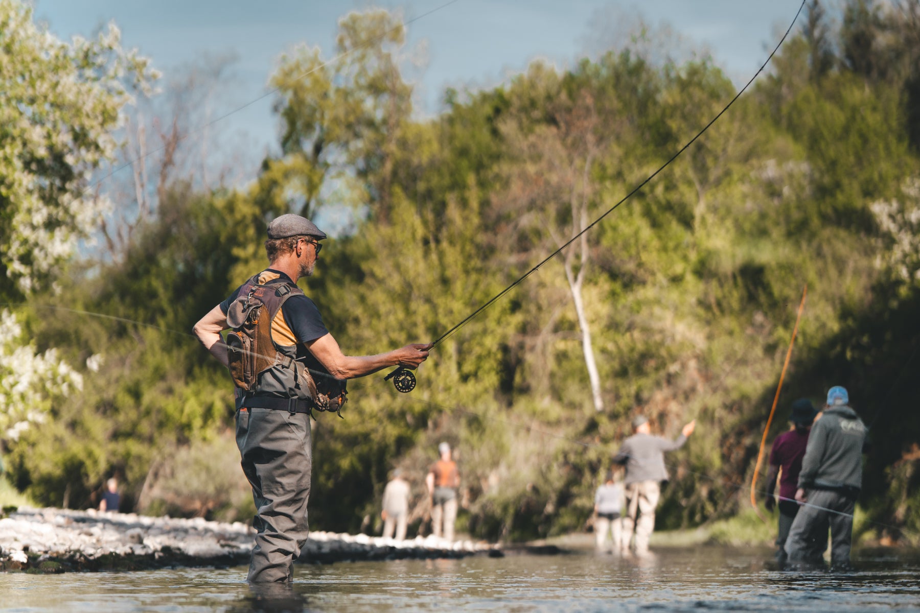 Fortgeschrittener Fliegenfischer im Drill am Schweizer Fluss, weitere Teilnehmer im Hintergrund – Fortgeschrittenenkurs von Flyfishing Guide Service. Flyfishing Guide Service, Flyfish Bünti, Fliegenfischen mit Rolf Bünter, Fliegenfischen Schweiz, Fortgeschrittenenkurs, Fliegenfischer im Drill, Wurfweste, Schweizer Fluss, Fortgeschrittene Wurftechnik, Stammrevier Töss, fortgeschrittene Teilnehmer, Drill am Fluss, Fliegenfischer-Profi, Wurfweite, taktisches Fischen, Fortgeschrittenen-Coaching, Schweizer Aueng